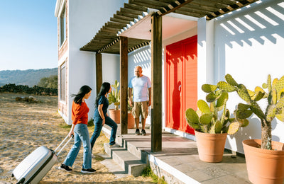 Two women with a suitcase walking up steps to a house with a man standing at the door.